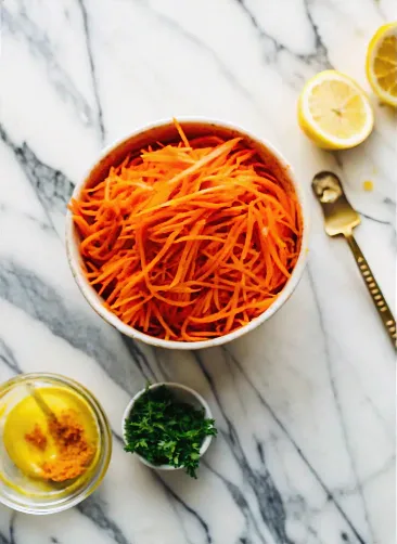 Shredded carrots in a bowl with lemon halves, mustard, and chopped parsley on a marble surface.