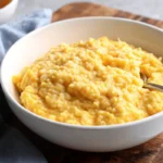 A bowl of creamy, cheesy rice with a silver spoon, served on a wooden board with a blue napkin.