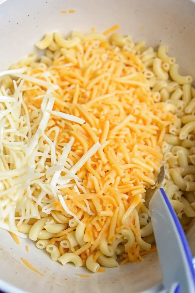 Shredded mozzarella and cheddar cheese added to cooked macaroni in a bowl for Mac and Cheese Meatloaf Casserole.