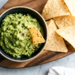 A bowl of chunky guacamole with a tortilla chip dipped in, surrounded by additional chips on a wooden platter.