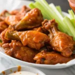 Buffalo wings being drenched in spicy sauce, with celery sticks on the side.