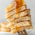 A close-up of a fork holding a bite-sized stack of fluffy buttermilk pancakes, drizzled with golden syrup.